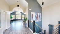 Located at 7001 W Cross Creek Trail in Brecksville, Ohio, this two-floor community space offers a generous and well-appointed layout across two distinct levels. The main floor greets visitors with a striking hallway and staircase area defined by soaring vaulted ceilings, bold slate-blue accent walls, rich wood-toned luxury vinyl plank flooring, and a sleek modern staircase featuring black metal railings with warm wood handrails. Large-scale artwork and pendant lighting add a sophisticated touch to the space. The adjacent living and dining area is bathed in natural light from expansive windows, including a dramatic arched window that frames outdoor views of lush, colorful foliage. Vaulted ceilings and deep blue walls lend this room an airy, refined ambiance with bistro-style seating for casual gatherings. A separate living room offers a cozy fireplace, crown molding, patterned carpet, and ample seating for relaxed conversation. The kitchen is outfitted with gray shaker-style cabinetry, white countertops, stainless steel appliances, and black hardware accents. The lower level houses a well-equipped exercise room featuring treadmills, ellipticals, free weights, an adjustable bench, floor-to-ceiling mirrors, rubber flooring, and access to restroom facilities — providing a comprehensive fitness experience within the property.