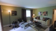 This single-floor living space is designed with a cohesive color palette centered around sage green walls, which lend a calm, earthy tone throughout the room. The living area is furnished with a cushioned sofa adorned with decorative pillows in complementary brown and tan tones, alongside a patterned armchair and matching ottoman that add visual interest and comfort. Dark brown curtain panels frame a central window, allowing natural light to filter into the space while providing contrast against the green walls. A classic table lamp with a warm-toned shade provides ambient lighting near the seating area. The room features a rectangular coffee table positioned centrally, topped with decorative accents including a bowl centerpiece. Wall art in abstract and earthy tones is thoughtfully placed on opposing walls, reinforcing the room's coordinated aesthetic. A patterned area rug in neutral and muted tones anchors the seating arrangement, while the surrounding flooring provides a clean contrast. A small side table with additional décor completes the arrangement, contributing to the room's organized and intentional design. Overall, the space presents a comfortable and well-appointed living environment.