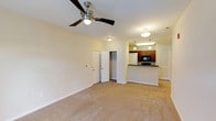 Located at 1001 Hayes Ln in Holly Springs, NC, this single-floor residence offers a practical and comfortable layout designed for everyday living. The open living area is generously proportioned, finished with wall-to-wall neutral carpeting and warmed by a ceiling fan overhead. A built-in closet provides convenient storage directly off the main living space. The living area flows seamlessly into the kitchen, which is defined by a raised breakfast bar with a dark countertop — ideal for casual dining or entertaining. The galley-style kitchen is well-equipped with rich medium-toned wood cabinetry, dark countertops, a double stainless steel sink, and black appliances including a refrigerator, range, and microwave. Light tile flooring and pendant lighting complete the kitchen's functional design. The bedroom is a calm, carpeted retreat featuring a ceiling fan and a window that allows natural light to filter in, creating a bright and airy atmosphere. The bathroom includes a combined tub and shower unit with built-in shelving, tile flooring, and a clean, neutral finish throughout. The space is characterized by a consistent warm-toned color palette, white trim work, and a straightforward layout that maximizes usability.