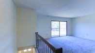 This well-maintained space offers a clean, open layout ideal for comfortable living. The main living area is finished with plush wall-to-wall carpeting in a cool, neutral tone, creating a warm and inviting atmosphere throughout. Smooth, light-painted walls complement the bright, airy feel of the space.

A prominent sliding glass door serves as a central architectural feature, flooding the room with natural light and providing direct access to an outdoor balcony. Views of mature trees and greenery beyond the balcony bring a sense of nature and tranquility into the interior.

A staircase near the entry features a classic wood handrail paired with dark metal balusters, adding a traditional design touch to the space. The stairwell is accented with small step lights, enhancing both safety and visual appeal. The smooth, clean ceiling throughout the living area reinforces the spacious, uncluttered feel of the interior.

Overall, the space presents a bright, functional, and inviting environment, characterized by its generous natural light, connection to the outdoors, and timeless design details.