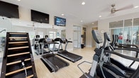 This fitness center is a comprehensively outfitted facility designed to support a full range of workout routines. The space is organized into distinct functional zones, each dedicated to a specific type of exercise. The cardio area houses multiple treadmills and elliptical machines, complemented by wall-mounted flat-screen televisions and ceiling fans that contribute to a comfortable workout environment. Full-length mirrors line the walls in several sections, visually expanding the space and allowing users to monitor their form. A dedicated cycling area features several stationary spin bikes positioned along a row of windows, which allow ample natural light to filter in and offer views of the surrounding outdoor landscape. The strength training section is generously equipped with a variety of cable and weight machines, including multi-functional gym stations and benches, alongside a vertical climbing ladder for functional fitness training. A dumbbell rack provides additional free-weight options. Light wood-style flooring runs throughout, lending a clean, contemporary aesthetic. Recessed lighting, ceiling fans, and large windows with roller shades maintain a bright and airy ambiance. Decorative wall accents and a clock add subtle character to the overall design, making this a polished and practical fitness facility.