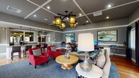 This thoughtfully designed single-floor clubhouse lounge blends comfort and functionality in an open, welcoming layout. The main lounge area makes an immediate impression with its dramatic dark coffered ceiling, adorned with a striking cluster of amber-glass pendant lanterns that cast warm, inviting light throughout the space. Rich hardwood flooring anchors the room, complementing the cool gray wall tones and crisp white wainscoting and trim.

The seating area is generously appointed with a large sectional sofa accented by teal and charcoal throw pillows, bold red accent chairs, and a central wooden coffee table — creating an inviting arrangement suited for both relaxed gatherings and casual conversation. Contemporary artwork in cool blues and greens punctuates the walls, adding color and sophistication.

Along one wall, a built-in workstation with white cabinetry and a granite-style countertop provides a dedicated area for computer use, ideal for productivity. Multiple windows dressed with dark curtain panels bring in natural light while offering a view to the outdoors.

Adjacent to the lounge, a bar-height counter with leather stools serves as a social hub, with a mirrored back wall amplifying the sense of space. The attached kitchenette features tile flooring, white cabinetry, dark countertops, and appliances including a coffee station and refrigerator, ensuring convenient refreshment access throughout the space.