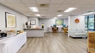 Located at 1099 N McMullen Booth Rd in Clearwater, Florida, this single-floor commercial space offers a thoughtfully designed, open-concept layout that balances functionality with a warm, inviting atmosphere. Light wood-look luxury vinyl flooring runs throughout, complementing the soft gray walls and drop-panel ceiling fitted with recessed fluorescent lighting.

The space is organized into several distinct functional zones. A comfortable lounge area features a mix of upholstered sofas, wicker armchairs, and accent tables arranged around a wall-mounted flat-screen television, with large floor-to-ceiling windows along the perimeter flooding the area with natural light and offering views of the surrounding greenery. A dedicated workstation zone includes a long white built-in desk with ample drawer storage and multiple computer monitors, anchored by a vibrant framed world map that serves as a focal point on the wall.

A refreshment station with a white countertop peninsula provides space for beverages and light food service, complete with a coffee machine, beverage dispenser, and display of fresh fruit. Near the entrance, a set of elegant white French doors with glass sidelights adds a refined architectural detail and enhances the sense of openness. The overall ambiance is clean, professional, and welcoming, making the space well-suited for client-facing or community-oriented use.