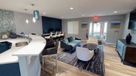 This single-floor community amenity space is thoughtfully designed with a coastal-inspired aesthetic, blending function and style throughout. The open layout flows seamlessly between distinct zones, all unified by light wood-look flooring, soft gray walls, and deep navy blue accent elements.

The centerpiece of the space is a spacious open kitchen featuring bold navy shaker-style cabinetry topped with crisp white countertops. A striking teal herringbone mosaic tile backsplash extends to the ceiling, adding a dynamic visual focal point. The kitchen is equipped with a built-in cooktop, stainless steel range hood, a large double sink, and a dedicated refreshment area with a full-size refrigerator and single-serve coffee station.

A curved white countertop bar with seating connects the kitchen to the main lounge area, providing a casual gathering spot. The lounge is furnished with a plush teal velvet sofa, modern accent chairs, and a geometric coffee table, all anchored by a navy abstract area rug. A wall-mounted flat-screen television and a sleek media console make this area ideal for entertainment.

A dedicated workstation zone along one wall features multiple computer monitors and high-top seating, catering to productivity needs. Natural light filters in through multiple windows and a glass-panel exterior door, which opens to an outdoor walkway. Coastal landscape artwork adorns the walls, reinforcing the space's relaxed, inviting ambiance.
