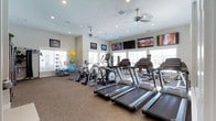 This fitness center offers a comprehensive workout environment designed to accommodate a variety of exercise routines. The single-story space is filled with natural light streaming through multiple windows fitted with white plantation shutters, creating a bright and welcoming atmosphere. Recessed lighting and dual ceiling fans ensure the room remains well-lit and comfortably ventilated during workouts.

The cardio section is prominently featured, housing a row of treadmills and elliptical trainers, along with additional cardio equipment toward the back of the room. Multiple flat-screen televisions are mounted along the walls, providing entertainment options for users during their sessions. The flooring consists of neutral-toned carpet, offering a practical surface throughout the space.

Along the perimeter, a dedicated stretching and functional training area is visible, equipped with a rack of colorful stability and medicine balls, as well as resistance training accessories. Motivational and decorative framed artwork adorns the walls, contributing to an energetic yet focused ambiance. Overall, the space is thoughtfully organized to support both cardiovascular and functional fitness training.