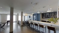 This single-floor space showcases a well-designed open-plan layout that seamlessly integrates multiple functional zones. A prominent white bar counter runs along one side of the space, lined with sleek, dark-upholstered bar stools that offer casual seating. Pendant lights hang above the counter, providing focused illumination and contributing to the modern aesthetic.

The space features a striking accent wall in deep blue-grey, adding depth and contrast against the predominantly white and neutral color palette. Built-in shelving is incorporated into this feature wall, offering both display and storage functionality. Adjacent to the bar area, a lounge or dining zone is defined by a patterned area rug, with contemporary dark chairs arranged around a table.

Structural columns are evenly spaced throughout the space, complementing the clean architectural lines. Large windows along the perimeter allow generous natural light to fill the interior, enhancing the open and airy feel. The polished concrete-style flooring runs continuously throughout, unifying the different zones. Recessed and pendant lighting ensure the space is well-lit throughout the day and evening, making it suitable for both social gatherings and relaxed use.