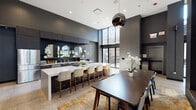 Located on the 14th floor of 19 S La Salle Street in Chicago's Loop, this sophisticated amenity space is defined by a bold, contemporary aesthetic characterized by dark charcoal walls, large-format stone tile flooring, and soaring double-height ceilings that lend a dramatic sense of volume throughout. The open-plan layout connects a fully equipped kitchen, a relaxed lounge, and a formal dining area in one cohesive environment.

The kitchen is outfitted with sleek dark wood cabinetry, white quartz countertops, stainless steel appliances including a French door refrigerator and built-in microwave, and a dark subway tile backsplash accented by arched mirrors. A generous island with bar seating serves as a central gathering point, illuminated by a polished globe pendant fixture.

The lounge area features plush sectional seating arranged around a linear fireplace set into a floor-to-ceiling stone accent wall, with a wall-mounted display screen above. Natural light pours in through floor-to-ceiling windows, with sliding glass doors providing direct access to an outdoor terrace furnished with lush greenery. An elevator lobby with seating completes this refined, amenity-rich floor.