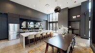 Located on the 14th floor of 19 S La Salle Street in Chicago's Loop, this sophisticated amenity space is defined by a bold, contemporary aesthetic characterized by dark charcoal walls, large-format stone tile flooring, and soaring double-height ceilings that lend a dramatic sense of volume throughout. The open-plan layout connects a fully equipped kitchen, a relaxed lounge, and a formal dining area in one cohesive environment.

The kitchen is outfitted with sleek dark wood cabinetry, white quartz countertops, stainless steel appliances including a French door refrigerator and built-in microwave, and a dark subway tile backsplash accented by arched mirrors. A generous island with bar seating serves as a central gathering point, illuminated by a polished globe pendant fixture.

The lounge area features plush sectional seating arranged around a linear fireplace set into a floor-to-ceiling stone accent wall, with a wall-mounted display screen above. Natural light pours in through floor-to-ceiling windows, with sliding glass doors providing direct access to an outdoor terrace furnished with lush greenery. An elevator lobby with seating completes this refined, amenity-rich floor.