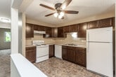 This single-story unit is designed with a functional, open layout that connects the living and kitchen areas through a distinctive pass-through opening. The kitchen is well-appointed with rich, dark espresso-toned cabinetry providing ample storage, light-colored countertops, a double-basin stainless steel sink, a dishwasher, a white electric range, and a full-size refrigerator. Stone-look tile flooring runs throughout the kitchen, adding durability and visual interest. A ceiling fan with a light fixture overhead enhances air circulation within the kitchen space.

The living area is generously sized, finished with neutral carpet, and features a sliding glass door flanked by vertical blinds, offering access to an outdoor area and bringing in natural light. A wall-mounted air conditioning unit and baseboard heating ensure year-round comfort.

The bedrooms are carpeted and naturally lit through large windows overlooking mature trees, each equipped with ceiling fans or overhead lighting and baseboard heaters. The bathroom includes a full tub-shower combination, a vanity with light countertops, warm wood cabinetry, and stone-look tile flooring. Textured ceilings are consistent throughout, and the overall color palette of warm beige tones gives the space a cohesive, neutral ambiance.