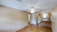 This single-story space offers a straightforward, functional layout with a warm, neutral aesthetic throughout. The main living area features wood-style hard flooring that extends across the room, complementing the beige-toned walls and white trim. A ceiling fan with an integrated light fixture is centrally mounted, providing both ventilation and ambient lighting beneath a textured popcorn ceiling. A vent in the ceiling suggests a central air system is in place.

The open layout connects the living area directly to the kitchen, which is visible along the right side of the space. The kitchen includes a refrigerator, additional white appliances, and what appears to be a countertop with a darker surface. A window in the kitchen area allows natural light to filter into the space.

Toward the back of the living area, a white interior door leads to an adjacent room or hallway, with an additional door visible beyond it, suggesting further connected rooms. The overall ambiance is clean and understated, with a simple, move-in-ready feel suited for practical, everyday living.