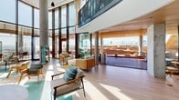 Located at 1515 Surf Ave in Coney Island, Brooklyn, this rooftop amenity space offers an exceptional outdoor and indoor retreat with sweeping views of the Atlantic Ocean and the landmark Parachute Jump. The multi-level layout is anchored by a resort-style pool, finished in bright aqua tile and lined with depth markings, accessible via wide entry steps with stainless steel railings. Adjacent to the pool, a generous sun deck is decked in warm hardwood and furnished with rows of modern white lounge chairs, perfect for sunbathing beneath open skies. A sculptural staircase with glass railings and a rich wood-paneled underside connects the deck levels, adding an architectural focal point to the outdoor space. A covered lounge area, sheltered beneath a curved wood-clad ceiling supported by a concrete column, provides a sophisticated seating nook that frames dramatic views of the Coney Island boardwalk and ocean beyond through floor-to-ceiling glass. Indoors, a bright interior lounge features soaring ceilings, expansive glazed walls that flood the space with natural light, and curated contemporary furnishings. A sleek kitchenette with light wood cabinetry and integrated appliances serves the interior lounge area. Lush potted plantings and glass perimeter railings complete this refined, resort-inspired amenity space.
