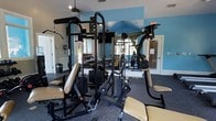 Located at 2155 Benton Blvd in Savannah, Georgia, this fitness center offers a dedicated, single-floor workout facility designed to support a well-rounded exercise routine. The space is finished with durable rubber flooring well-suited for both strength training and general fitness activities. A vibrant teal accent wall adds a lively, energizing atmosphere to the room, complemented by large mirrors that line the walls to enhance the sense of openness and allow users to monitor their form during workouts. The centerpiece of the facility is a multi-station cable and strength machine, providing a comprehensive range of resistance training options including cable pulldowns, chest presses, and leg extensions. Additional benches and adjustable seating stations round out the strength training offerings. Natural light filters into the space through windows, keeping the environment bright and welcoming, while a ceiling fan supports air circulation during intensive sessions. The room's layout is thoughtfully organized to allow users to move freely between equipment. White-paneled doors provide clean transitions between areas. Overall, this fitness center delivers a functional, well-equipped environment suitable for residents or members seeking convenient access to quality exercise amenities in the Savannah area.