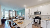This single-floor residence showcases a thoughtfully designed open-concept layout that seamlessly connects the kitchen, dining, and living areas. The living room is defined by a signature teal accent wall, wood-look flooring, and abundant natural light streaming through large windows dressed with blinds and warm-toned drape panels. A ceiling fan with an integrated light fixture anchors the space, while gold-toned sconce lighting adds a refined touch. A compact dining area with a round table sits adjacent to the front entry door, making efficient use of the layout.

The kitchen is well-equipped with white shaker-style cabinetry, granite countertops, stainless steel appliances including a range, microwave, and refrigerator, and a central island with a built-in sink and bar seating. Recessed lighting illuminates the work surfaces throughout.

The bedroom offers a ceiling fan, wood-look flooring, and a large window with outdoor views, creating a calm and restful atmosphere. The bathroom features a granite-topped vanity, undermount sink, and wood-look flooring, maintaining the consistent finish quality found throughout the residence.