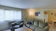 This single-floor residential space offers a comfortable and functional layout centered around a welcoming living area. The living room is furnished with a plush, earth-toned sofa and matching armchair, creating an inviting seating arrangement ideal for relaxation. A mosaic-tiled coffee table anchors the space and adds a decorative touch to the room's casual aesthetic. Natural light filters generously through large vertical blinds, brightening the room and contributing to its open, airy feel. A window air conditioning unit is installed beneath the window, ensuring year-round comfort. Warm ambient lighting is provided by a tall floor lamp positioned in the corner, casting a soft glow across the neutral-toned walls. A vibrant cityscape wall print serves as a focal point, adding a modern and artistic element to the otherwise understated décor. The textured ceiling and smooth walls maintain a clean, simple architectural character throughout. The space also features a cat tree near the window, reflecting a pet-friendly environment. Overall, the living area strikes a balance between practicality and comfort, making it a functional and pleasant everyday living space.