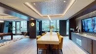 This single-floor amenity space is thoughtfully designed to offer multiple distinct zones for socializing, entertainment, and collaboration. The main gathering area centers around a substantial marble-topped communal table surrounded by mustard-yellow upholstered chairs, positioned beneath a striking coffered ceiling with a textured metallic inlay and a sleek linear pendant light. Floor-to-ceiling windows flood this area with natural light and offer views of a lush outdoor courtyard. Adjacent to the table, a built-in credenza with cabinetry and a large wall-mounted display screen provides functional utility for presentations or media viewing.

The entertainment zone features a full-sized billiards table set atop a bold patterned area rug, flanked by tufted leather banquette seating and curated abstract artwork. A gold-toned coffered ceiling with a circular pendant adds warmth and visual drama to this space.

A more intimate lounge nook offers a cozy retreat, furnished with a plush velvet armchair in deep yellow, a burgundy sofa, and a vibrant pink wave-patterned rug beneath a richly textured dark ceiling with ambient LED lighting.

The entryway corridor connects these areas seamlessly, featuring dark marble tile flooring, framed artwork, and refined architectural detailing, establishing an elevated tone throughout the entire space.