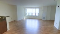 This single-floor space presents a clean, minimalist aesthetic designed to maximize both comfort and functionality. The open layout provides a generous sense of space, anchored by smooth light-toned hardwood flooring that runs throughout and reflects natural light beautifully. The walls are finished in crisp white, lending the interior a fresh and airy ambiance that enhances the sense of openness.

A prominent multi-pane window serves as the visual centerpiece of the main living area, allowing an abundance of natural light to pour in and offering views of the surrounding exterior. The window's wide span ensures the space remains bright throughout the day, creating an inviting and comfortable atmosphere.

The room features recessed ceiling lights for additional illumination during evening hours, complementing the natural daylight. A wall-mounted HVAC vent is neatly integrated into the design, ensuring climate comfort without compromising the clean lines of the interior. The white baseboards and trim provide a subtle but polished finishing touch throughout.

Overall, the space delivers a practical yet welcoming environment, well-suited for a variety of uses. Its straightforward, uncluttered design allows for flexible furnishing and personalization, making it an excellent canvas for any occupant looking to create a functional and comfortable living or working environment.