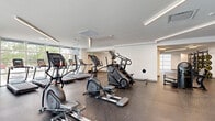 This expansive fitness center offers a comprehensive, well-equipped exercise facility spread across a single, thoughtfully designed floor. The space is organized into distinct functional zones, each catering to different training disciplines.

The cardio area features a row of treadmills and elliptical machines positioned along large floor-to-ceiling windows, flooding the space with natural light and offering views to the outdoors. Stair climbers round out the cardio offerings. A dedicated strength training zone houses a variety of selectorized weight machines, power racks, and free-weight benches, complemented by a striking motivational mural that anchors the room with bold fitness-inspired graphics and typography.

A separate cycling studio provides a polished hardwood floor, Peloton-style spin bikes, and a full-length wall mirror — ideal for group classes or individual sessions. The free weight and functional training area includes squat racks, plate-loaded barbells, adjustable benches, and open floor space with rubber matting for dynamic movements.

Throughout the facility, clean white walls and linear LED lighting create a bright, energizing atmosphere. Durable rubber flooring covers the equipment zones, providing safety and sound absorption. Wide, well-lit corridors connect each area seamlessly, contributing to an open, uncluttered layout that supports an effective and comfortable workout experience.