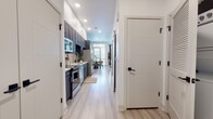 This thoughtfully designed single-floor residence combines modern aesthetics with functional living. Light wood-tone flooring flows continuously throughout, unifying the space with a clean, contemporary feel.

The galley-style kitchen is a standout feature, outfitted with dark charcoal shaker cabinets extending to the ceiling for generous storage, a white tile backsplash, light countertops, and a full suite of stainless steel appliances including a range, dishwasher, over-the-range microwave, and a French door refrigerator with a water and ice dispenser.

A wide hallway connects the kitchen to the dining and living areas, flanked by shiplap-paneled doors with matte black hardware that add a modern farmhouse touch. The dining area accommodates a round table with cushioned chairs and is brightened by a large window with access to natural light. The living room offers a comfortable, open layout with large windows that invite further natural light.

The bedroom features soft gray walls, recessed lighting, and ample space for a queen or king bed. The bathroom includes hexagonal tile accents, dark vanity cabinetry, and decorative wall art. A stacked washer and dryer unit is conveniently located nearby, completing this well-rounded residence.