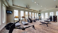 Located at 240 Park Place Blvd in Waxahachie, TX, this fitness center is a comprehensively outfitted facility designed to support a full range of workout routines. The main exercise floor is generously sized, featuring patterned carpet flooring and tray ceilings accented with recessed lighting and ceiling fans that promote air circulation throughout. Large windows line the walls, flooding the space with abundant natural light and offering views of the outdoor pool area and surrounding community. The cardio section is well-stocked with treadmills, stair climbers, ellipticals, stationary bikes including a Peloton, and a Concept2 rowing machine. Mounted flat-screen TVs are positioned throughout for entertainment during workouts. The strength training area includes a functional cable trainer, a Smith machine, adjustable benches, and a dumbbell rack, all reflected in floor-to-ceiling mirrors that enhance the sense of space. A dedicated studio room features hardwood-style flooring, wall-mounted mirrors, a mounted TV, medicine ball racks, TRX suspension trainers, step platforms, and agility equipment — ideal for functional and group-style training. Together, these areas create a professional and versatile fitness environment suited to a wide variety of fitness goals.