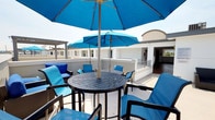 This rooftop terrace is a well-appointed outdoor amenity space designed for both relaxation and entertaining. The terrace is finished in crisp white stucco walls and a smooth, light-toned deck surface that reflects the abundant natural sunlight throughout the day. Vibrant blue patio umbrellas are positioned throughout the space, providing generous shade over multiple seating configurations, including round slatted dining tables paired with blue-cushioned chairs and wicker lounge seating accented with coordinating blue throw pillows. A pergola structure anchors one section of the terrace, adding architectural interest and an additional covered gathering area. The outdoor kitchen is a standout feature, equipped with stainless steel built-in grills set into a clean stone-topped counter, ideal for alfresco dining and social gatherings. Sleek modular black lounge chairs are positioned nearby, creating a casual conversation area. The terrace is enclosed by low white parapet walls, beyond which expansive open views of the surrounding neighborhood unfold, including palm trees, mature greenery, and distant mountain silhouettes visible on clear days. The overall ambiance is bright, airy, and inviting, making this rooftop terrace a versatile outdoor retreat suited for both casual relaxation and lively social events.