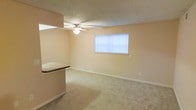This single-floor space features an open living area finished with wall-to-wall carpet in a neutral tone, creating a clean and functional environment. The room is illuminated by a ceiling fan with an integrated light fixture, as well as natural light filtering through a window fitted with horizontal blinds. The walls are painted in a soft beige, complementing the light-colored carpet and white baseboards throughout. A partial countertop divider, finished with a dark laminate surface, separates the living area from an adjacent space, adding a subtle architectural element to the otherwise open layout. The textured ceiling adds a traditional finish consistent with the overall design. The space presents a straightforward, practical layout with ample open floor space suitable for a variety of furniture arrangements.