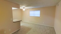 This single-floor space features an open living area finished with wall-to-wall carpet in a neutral tone, creating a clean and functional environment. The room is illuminated by a ceiling fan with an integrated light fixture, as well as natural light filtering through a window fitted with horizontal blinds. The walls are painted in a soft beige, complementing the light-colored carpet and white baseboards throughout. A partial countertop divider, finished with a dark laminate surface, separates the living area from an adjacent space, adding a subtle architectural element to the otherwise open layout. The textured ceiling adds a traditional finish consistent with the overall design. The space presents a straightforward, practical layout with ample open floor space suitable for a variety of furniture arrangements.