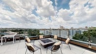 This rooftop terrace offers an elevated outdoor retreat with a thoughtfully designed layout centered around a sleek, square fire pit finished in a warm bronze tone and filled with dark fire glass. Surrounding the fire pit is a curated arrangement of modern metal mesh lounge chairs, each accented with plush, patterned throw pillows, creating a welcoming and social atmosphere. The terrace floor is finished with large-format light-toned tiles that lend a clean, contemporary aesthetic to the space. Glass panel railings line the perimeter, preserving unobstructed, panoramic views of the city skyline while maintaining an open, airy feel. The surrounding urban landscape is visible in all directions, with a mix of mid-rise buildings, greenery, and landmark architecture visible on the horizon. The open-sky exposure ensures abundant natural light throughout the day, making this terrace an ideal setting for both daytime relaxation and evening gatherings. The combination of the fire pit, modern furnishings, and sweeping cityscape views makes this rooftop an exceptional outdoor amenity space suited for entertaining or unwinding in an elevated urban environment.