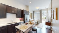 Located within the Riverwalk community in Plainsboro, NJ, this single-floor residence combines modern design with functional living. The open-concept layout seamlessly connects the kitchen and living area, creating a cohesive and airy environment enhanced by notably high ceilings throughout.

The kitchen is outfitted with rich dark-wood cabinetry, light quartz countertops, a classic white subway tile backsplash, and stainless steel appliances including a refrigerator, range, microwave, and dishwasher. A freestanding island with a marble-style surface provides additional prep space and casual seating with bar stools. Warm hardwood-style flooring runs throughout the kitchen and living areas.

The living room is bright and welcoming, benefiting from oversized windows that invite abundant natural light. A sliding glass door opens directly to a private balcony with exterior views of the surrounding community. Tall ceilings amplify the sense of space.

The bedroom is a calm, neutral retreat featuring large windows dressed with patterned curtains and wood blinds, offering excellent light control. The bathroom features large-format marble-look tile flooring, a dark-wood double vanity with an illuminated mirror, and an adjacent soaking tub visible through a connecting doorway.