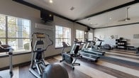 Located at 10 Kenley Way in Birmingham, Alabama, this fitness center is a thoughtfully designed single-floor facility built to support a well-rounded workout experience. The space features vaulted ceilings with recessed lighting that create an open, airy atmosphere, while large windows allow natural light to pour in and offer views of the surrounding exterior. Wide-plank hardwood flooring runs throughout, lending a clean, contemporary aesthetic.

The cardio zone is outfitted with multiple treadmills, an elliptical trainer, and a stationary bike, all positioned to take advantage of the natural light and window views. Full-length mirrored walls expand the sense of space and allow users to monitor their form during workouts. A wall-mounted television provides entertainment options for cardio sessions, and a dedicated split-system air conditioning unit ensures comfortable climate control.

The strength training area includes a multi-function cable and weight machine suitable for a wide range of exercises, complemented by a free weight rack with dumbbells. A tufted black leather bench and open dark-framed shelving add a modern lounge-like element, while geometric metallic wall art panels introduce a refined, stylish touch. French doors provide access to an adjacent outdoor area, enhancing connectivity and ventilation throughout the space.