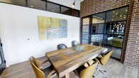 This single-floor meeting space combines industrial and contemporary design elements to create a functional yet visually engaging environment. At its center sits a substantial reclaimed wood conference table, its rich, natural grain providing a warm focal point surrounded by a mix of leather and upholstered chairs in complementary tan and dark tones. The seating arrangement comfortably accommodates multiple attendees, making the room well-suited for collaborative meetings or presentations.

Exposed brick detailing along one wall adds texture and an urban, industrial character to the space, while crisp white walls provide contrast and visual balance. A large abstract artwork in muted yellows and blues anchors one wall, contributing a creative and professional atmosphere. Overhead, a simple pendant light fixture provides focused illumination, complementing the natural light that filters through the large black-framed glass doors and transom windows. These doors connect the meeting room to an adjacent area, which appears to feature a bar or lounge setup, hinting at the broader mixed-use nature of the overall space. Light hardwood flooring ties the room together with a clean, modern finish.