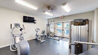 Located at 1601 Cottleville Pkwy in Cottleville, Missouri, this fitness center offers a comprehensive workout environment within a compact, efficiently designed single-room layout. The space features durable rubber flooring throughout, providing a safe and functional surface for all types of exercise.

The cardio section is equipped with treadmills, an elliptical trainer, and a stationary recumbent bike, while a wall-mounted flat-screen TV keeps users entertained during workouts. A multi-function cable machine and a dedicated weight area round out the strength training offerings. The free weight zone includes a full rack of dumbbells and medicine balls, a weight bench, and kettlebells, complemented by a large wall mirror that enhances the sense of space and assists with form.

Natural light filters into the fitness center through several windows and a set of glass French doors that open directly to an outdoor pool area, creating a bright and welcoming atmosphere. The neutral-toned walls and overhead lighting maintain a clean, focused environment. A ceiling fan provides additional air circulation, and the space is designed to support a well-rounded fitness routine in a community-centered setting.