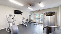 Located at 1601 Cottleville Pkwy in Cottleville, Missouri, this fitness center offers a comprehensive workout environment within a compact, efficiently designed single-room layout. The space features durable rubber flooring throughout, providing a safe and functional surface for all types of exercise.

The cardio section is equipped with treadmills, an elliptical trainer, and a stationary recumbent bike, while a wall-mounted flat-screen TV keeps users entertained during workouts. A multi-function cable machine and a dedicated weight area round out the strength training offerings. The free weight zone includes a full rack of dumbbells and medicine balls, a weight bench, and kettlebells, complemented by a large wall mirror that enhances the sense of space and assists with form.

Natural light filters into the fitness center through several windows and a set of glass French doors that open directly to an outdoor pool area, creating a bright and welcoming atmosphere. The neutral-toned walls and overhead lighting maintain a clean, focused environment. A ceiling fan provides additional air circulation, and the space is designed to support a well-rounded fitness routine in a community-centered setting.