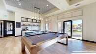 This contemporary single-floor amenity space is thoughtfully designed to serve as a social and recreational hub. The main gathering area is anchored by a full-size billiards table set beneath a striking tray ceiling adorned with a bold geometric patterned inset, complemented by a modern globe pendant chandelier that draws the eye upward. Light wood-tone flooring runs throughout, lending warmth and continuity to the space.

The wet bar area is a standout feature, showcasing a herringbone tile backsplash, open display shelving, under-counter lighting, and a stainless steel sink — ideal for entertaining. Large windows and glass doors flood the space with natural light and provide direct views of and access to an outdoor area with patio seating.

A dedicated lounge nook offers a more intimate retreat, featuring a curved boucle sofa, curated wall art, dual circular mirrors, and a flat-screen TV mounted against a dark shiplap accent wall. Adjacent lounge seating includes a pair of mustard-toned barrel chairs arranged around a coffee table on a patterned area rug, perfect for casual conversation or relaxation.

The catering kitchen is sleek and functional, outfitted with slate-grey cabinetry, white quartz countertops, open shelving, a herringbone tile backsplash, and ample counter space on both sides — all bathed in natural light from a large exterior-facing window.