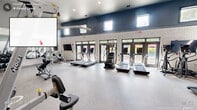 Located at 1400 E Cornwallis Rd in Durham, NC, this well-appointed fitness center offers a comprehensive exercise environment across a generously sized single-floor layout. The room features soaring ceilings accented by a bold navy upper wall, contrasted with white-painted brick below, creating a modern and energizing aesthetic. Natural light pours in through a row of large glass doors and elevated clerestory windows along multiple walls, offering views of the adjacent outdoor pool area and keeping the space feeling open and airy. The durable speckled rubber flooring provides both safety and comfort throughout. The cardio zone is well-stocked with multiple treadmills, stair climbers, elliptical machines, and stationary bikes, all positioned to take advantage of the outdoor views. The strength training area includes a full rack of dumbbells displayed along a floor-to-ceiling wall mirror, cable machines, weight benches, and adjustable resistance equipment. Additional functional fitness accessories such as medicine balls, stability balls, and resistance tools are neatly organized along the walls. A wall-mounted flat-screen television adds convenience for entertainment during workouts. Recessed lighting and a ceiling fan complete the space, ensuring comfort and visibility at all hours.