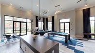 This single-floor amenity lounge is designed with a contemporary aesthetic, combining functionality and style in a spacious, open layout. Soaring ceilings create an airy, expansive atmosphere, while large floor-to-ceiling windows dressed in dramatic dark curtains flood the interior with natural light and offer views of the surrounding outdoor environment.

At the center of the space, a sleek billiards table anchored on a vibrant blue and white geometric-patterned area rug serves as the room's focal point, reinforcing the modern, energetic design theme. A substantial dark-toned counter or bar surface in the foreground adds a practical element, complemented by cabinetry below.

Overhead, an eclectic mix of pendant lighting fixtures — including globe and metallic disco-style pendants — adds visual interest alongside a ceiling fan that enhances comfort. The lounge area features contemporary upholstered seating in neutral and teal tones, creating inviting gathering spaces. Neutral gray walls and light flooring provide a clean backdrop that ties the bold design accents together, resulting in a polished, social atmosphere well-suited for recreation and relaxation.