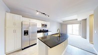 This well-appointed single-floor residence offers a thoughtfully designed open-concept layout that seamlessly connects the kitchen and living areas. The kitchen is a standout feature, showcasing crisp white cabinetry, striking dark granite countertops, and a suite of stainless steel appliances including a French door refrigerator with ice maker, electric range, and built-in microwave. A curved kitchen island with an undermount sink and brushed gold faucet serves as a functional centerpiece, providing additional prep space and a natural gathering point. Track lighting overhead illuminates the kitchen with precision, enhancing both the functionality and aesthetic of the space. The flooring transitions from clean, large-format tile in the kitchen to plush carpet in the living area, offering comfort underfoot. The living space is bathed in natural light, courtesy of a large sliding glass door that opens onto a private balcony, inviting the outdoors in. Neutral wall tones and a light, airy color palette throughout create a calm, welcoming ambiance, making this space an ideal blend of modern style and everyday practicality.