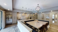 This single-floor space showcases a thoughtfully designed open-concept kitchen and dining area that seamlessly blends elegance with functionality. The kitchen is anchored by an expansive white island with a light granite countertop, providing generous workspace and serving as the room's focal point. A built-in banquette with deep navy tufted upholstering wraps around a large dining table, creating an inviting and intimate seating area integrated directly into the kitchen layout.

The cabinetry is finished in crisp white with clean lines, complemented by a white subway tile backsplash and under-cabinet lighting that enhances the bright, airy atmosphere. Overhead, a striking orb-style chandelier with a weathered wood and metal finish adds a rustic-chic contrast to the otherwise refined aesthetic, while a series of amber glass pendant lights hang in a coordinated row above the island.

The flooring features a geometric patterned tile design that adds visual interest and sophistication to the space. Recessed lighting throughout ensures even illumination, while the soft neutral color palette of whites, grays, and warm wood tones creates a cohesive and welcoming ambiance. A glass-paned door visible in the background hints at additional connected spaces beyond.