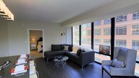 This contemporary single-floor residence offers a refined urban living experience defined by clean lines, modern finishes, and an abundance of natural light. The living area is an immediate highlight, featuring dramatic floor-to-ceiling windows that frame sweeping city views and flood the space with daylight. Dark hardwood flooring runs throughout, adding warmth and visual contrast against the neutral-toned walls and crisp white ceilings. The living room is thoughtfully arranged around a sectional sofa and a geometric glass coffee table, creating an inviting yet sophisticated atmosphere. A stylish arc floor lamp adds an elegant design touch, complementing the modern aesthetic of the space. The open layout allows for a seamless flow between the living area and the adjacent bedroom, which is visible through a connecting doorway. The bedroom continues the neutral palette with soft, layered bedding and coordinating furnishings. Throughout the space, the interplay of city light, contemporary décor, and carefully selected materials creates an atmosphere that is both functional and visually engaging — ideal for those seeking a modern urban lifestyle with a strong sense of style.