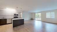 This single-floor residence offers a thoughtfully designed open-concept layout that seamlessly connects the kitchen and living area. The kitchen is a standout feature, equipped with stainless steel appliances including a refrigerator, range, microwave, and dishwasher. Sleek two-tone cabinetry — combining light grey uppers with rich dark wood lowers — pairs with a crisp white countertop and a mosaic tile backsplash. A central island with pendant lighting provides additional prep space and serves as a natural gathering point. Light wood-style flooring flows throughout the open living area, which is bathed in natural light from a large sliding glass door leading to a private balcony with views of mature trees and lush greenery. A secondary window further brightens the space. The bedroom features plush carpeting, a window overlooking the outdoors, and ample closet space. The bathroom is finished with warm-toned ceramic tile extending from floor to ceiling in the tub surround, complemented by coordinating floor tile, offering a clean and classic aesthetic. Overall, the residence delivers a modern, comfortable living environment with a neutral palette and functional design throughout.