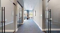 This contemporary single-floor space is designed with a polished, hospitality-inspired aesthetic that balances functionality with visual appeal. The entrance lobby immediately impresses with soaring floor-to-ceiling windows framed in dark metal, flooding the space with abundant natural light and offering clear views to the street outside. Exposed wood beam accents near the ceiling add warmth against the otherwise crisp, neutral palette of white and gray tones. A reception desk anchored near the entry sets a welcoming tone, while large-format tile flooring transitions seamlessly into patterned blue-toned carpet throughout the corridors and gathering areas.

A central highlight is the wet bar area, featuring dark cabinetry, built-in appliances, and a bold teal accent wall adorned with a graphic typographic mural. High-top seating with blue upholstered chairs provides an inviting social hub. Curated artwork is displayed throughout, including vibrant pop-art pieces and a striking cityscape canvas, contributing to a gallery-like atmosphere.

The corridor leads to enclosed private meeting rooms with glass-fronted walls and sleek black-framed sliding doors, offering privacy while maintaining visual openness. One meeting room features a vivid teal accent wall paired with plush orange velvet chairs around a dark conference table, creating a bold, energetic environment for focused collaboration.