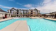 Located at 2700 Whakatane Ln in Fuquay-Varina, NC, this community clubhouse serves as a central gathering hub designed with upscale resort-style amenities. The exterior showcases a refined craftsman aesthetic, combining stacked stone column bases, board-and-batten siding, and decorative gabled rooflines that carry a cohesive architectural identity throughout the property.

The covered outdoor lounge is a standout feature, offering a welcoming transition between indoor and outdoor spaces. White French doors with transom windows provide entry into the interior, while the lounge itself is equipped with wicker and upholstered seating, a ceiling fan, and a striking stacked-stone wall anchored by a built-in gas fireplace and an outdoor-mounted television — ideal for year-round relaxation and entertainment.

Stately white columns set atop stone piers define the perimeter of the covered porch, which opens via a gated black iron fence directly to the resort-style pool deck. The expansive pool is surrounded by a generous sun deck lined with lounge chairs and umbrellas, offering sweeping views of the multi-story residential buildings and lush landscaping beyond. Palm trees accentuate the outdoor areas, reinforcing the leisure-oriented atmosphere of this well-appointed community amenity space.