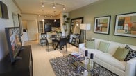 This single-floor residence showcases an open-concept layout that connects the kitchen, dining, and living areas in one cohesive, flowing space. The kitchen features dark cabinetry, black appliances, and a light-toned breakfast bar that serves as a natural transition into the dining area. Pendant lighting and track lighting illuminate the space, adding warmth and definition to each zone.

The dining area accommodates a round table with upholstered chairs, positioned atop a circular area rug, creating an intimate setting. A large framed mirror leans against the wall nearby, enhancing the sense of depth and space.

The living area is anchored by a white sofa dressed with sage green and patterned accent pillows, complementing the soft green accent wall behind it. A glass-top coffee table sits over a textured dark area rug, adding contrast to the light carpeted flooring. Decorative accessories, including candle holders and books, are neatly arranged throughout. A framed abstract artwork and a tall floor lamp complete the refined, contemporary aesthetic of this well-appointed space.