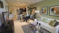 This single-floor residence showcases an open-concept layout that connects the kitchen, dining, and living areas in one cohesive, flowing space. The kitchen features dark cabinetry, black appliances, and a light-toned breakfast bar that serves as a natural transition into the dining area. Pendant lighting and track lighting illuminate the space, adding warmth and definition to each zone.

The dining area accommodates a round table with upholstered chairs, positioned atop a circular area rug, creating an intimate setting. A large framed mirror leans against the wall nearby, enhancing the sense of depth and space.

The living area is anchored by a white sofa dressed with sage green and patterned accent pillows, complementing the soft green accent wall behind it. A glass-top coffee table sits over a textured dark area rug, adding contrast to the light carpeted flooring. Decorative accessories, including candle holders and books, are neatly arranged throughout. A framed abstract artwork and a tall floor lamp complete the refined, contemporary aesthetic of this well-appointed space.