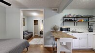 This efficiently designed one-bedroom, one-bathroom unit makes excellent use of its compact footprint across a single floor. The interior is finished with wood-look tile flooring throughout, soft sage-green walls, and textured ceilings that lend a consistent, cohesive aesthetic to the space.

The kitchen is a functional highlight, featuring white cabinetry, a light-toned countertop with a tile backsplash, and stainless steel appliances including a range, refrigerator, and microwave. Open wall-mounted shelving provides additional storage and display space. A freestanding butcher-block island with bar stools doubles as a dining area, maximizing the use of the open layout. A window near the entry allows natural light into the kitchen.

The bedroom is a comfortable retreat, accommodating a full-sized bed and offering a ceiling fan with a light fixture for year-round comfort. A window with blinds brings in natural light, and a dedicated closet with sliding doors provides ample storage. The room opens into a small adjacent living area with a sofa, expanding the functional space.

The bathroom is clean and practical, equipped with a glass-enclosed tiled stand-up shower, a pedestal sink, and a toilet. Hooks near the shower offer convenient towel storage. The overall layout reflects a smart, well-organized design suited for efficient single-occupant living.