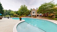 Located at 2951 Satellite Blvd in Duluth, Georgia, this amenity space offers a thoughtfully designed outdoor and indoor experience ideal for relaxation and social gatherings. At its centerpiece is a generously sized freeform swimming pool with a smooth concrete surround, lined with lounge chairs and vibrant blue market umbrellas. The pool area is framed by mature trees, ornamental grasses, colorful flowering shrubs, and tiered stone retaining walls, creating a lush, resort-like atmosphere with ample natural greenery.

A covered entrance porch features stone-clad columns, a ceiling fan, and bold orange seating, offering a welcoming transitional space between the outdoors and the interior. The indoor clubhouse kitchen is equipped with stainless steel cabinetry, granite-style countertops, a full-size stainless refrigerator, an under-counter refrigeration unit, and a wall-mounted flat-screen TV, all set against soft teal walls with crown molding.

The interior gathering area includes retro-style round bar tables with chrome barstools and a distinctive glass-panel garage door that opens fully to the outdoors, seamlessly blending the indoor and outdoor spaces. Patterned carpet flooring and track lighting complete the modern yet inviting aesthetic of this well-appointed community amenity space.