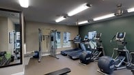 This fitness center offers a comprehensive workout environment designed to support a wide range of exercise routines. The space is finished with neutral-toned walls accented by a deep green feature wall, creating a focused and energizing atmosphere. Overhead fluorescent lighting ensures the room is well-lit throughout, complemented by natural light streaming in through multiple windows that offer views of the surrounding exterior.

The cardio section is prominently featured, housing multiple treadmills and an elliptical machine, all equipped with individual digital displays for tracking workout metrics. A multi-function cable weight machine anchors the strength training area, providing versatility for a variety of resistance exercises. A stability ball and additional free weights round out the available equipment, catering to both functional fitness and traditional strength training needs.

The flooring is finished in a durable, low-pile carpeted surface well-suited for a fitness environment. The ceiling-mounted fan aids in air circulation, keeping the space comfortable during use. The layout is thoughtfully arranged to allow ample room for movement between stations, making this fitness center a practical and well-appointed facility for daily exercise.