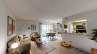 Located in Austin, TX, this one-bedroom, two-bathroom unit offers a thoughtfully arranged single-floor layout that maximizes every square foot. The open-concept living and dining area is anchored by light wood-style flooring, a textured ceiling, and a sliding glass door that floods the space with natural light while providing direct access to a private outdoor patio with a grassy area. The living room comfortably accommodates a full seating arrangement, complemented by wall art and an arc floor lamp that add warmth and character. Adjacent to the living space, the dining area features an open wood shelving unit that serves as both storage and a design element, further enhancing the connection to the patio. The kitchen is separated by a half-wall pass-through counter with granite-style countertops, white cabinetry, and a full-size refrigerator. The bedroom is carpeted and fitted with a ceiling fan, natural light from a window with blinds, and a dedicated closet. Two bathrooms complete the unit — one featuring a full tub/shower combination and the other a vanity with a granite-style countertop and framed mirror — providing practical convenience for residents.