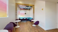 Located on Magnolia Boulevard in the North Hollywood neighborhood of Los Angeles, this single-floor commercial space serves as a café — identified by signage as Milano Cyber Cafe. The interior design strikes a balance between contemporary minimalism and artistic expression, creating an inviting atmosphere for guests.

The main seating area features warm wood-tone flooring throughout, crisp white walls, and high ceilings that lend a sense of openness. A distinctive architectural nook anchors one end of the space, framed by warm wood trim and outfitted with a teal upholstered L-shaped banquette. Above this nook, a dramatic ceiling mural depicting a vivid, moody cloudscape adds a bold artistic focal point, complemented by a textured pendant light hanging from its center.

Seating throughout the space consists of sleek, tulip-base round tables paired with modern plum-colored pod-style chairs, offering a cohesive and contemporary aesthetic. The entrance area showcases white paneled walls with a decorative geometric pattern, a glass-paned door, and windows fitted with gradient pink-to-yellow roller shades that diffuse soft, warm natural light into the room. The overall ambiance is modern, creative, and welcoming.
