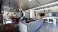 This single-floor space showcases a compelling blend of industrial and contemporary design, creating an inviting and visually dynamic environment. The open-plan lounge area is anchored by a large, tufted gray sectional sofa that serves as the centerpiece, complemented by rich leather armchairs and a vibrant floral-patterned area rug that adds a bold splash of color to the space. Exposed silver ductwork runs along the dark-painted ceiling, lending an industrial character that is elegantly balanced by a striking multi-arm chandelier and a system of track lighting that illuminates the room with precision. Pendant globe lights hang near the bar area, which features a sleek countertop, bar seating, and a wall-mounted flat-screen TV set against a textured tile accent wall — making it an ideal spot for socializing or casual viewing. The flooring transitions between large-format tile and the statement area rug, defining distinct zones within the open layout. Artwork adorns the walls, adding personality and a curated aesthetic throughout. The overall ambiance is modern and urban, with thoughtful design details that balance comfort and style, making this space well-suited for gathering, relaxing, or entertaining.