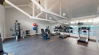 This fitness center is a thoughtfully designed single-floor space that combines architectural character with practical functionality. The room is defined by its striking vaulted ceiling, featuring exposed white structural trusses and white-painted wood paneling that lend the space an open, airy feel. Pendant globe lights hang from the ceiling, providing warm, even illumination throughout.

The walls are finished in painted white brick, contributing to the clean and bright aesthetic. A large full-wall mirror runs along one side of the room, visually expanding the space and allowing users to monitor their form during workouts. Large sliding glass doors and clerestory windows along one wall flood the interior with natural light and offer views of an outdoor pool area, creating a seamless connection to the exterior.

The fitness floor is covered in durable dark rubberized flooring with clearly marked orange spacing indicators. Equipment includes multiple treadmills, an elliptical trainer, stationary bikes, a multi-station cable machine, a weight bench, and a medicine ball rack stocked with various sizes. One wall features an eye-catching motivational mural composed of fitness-themed words in bold typography, adding energy and inspiration to the environment. A wall-mounted television provides additional entertainment or instructional content during workouts.